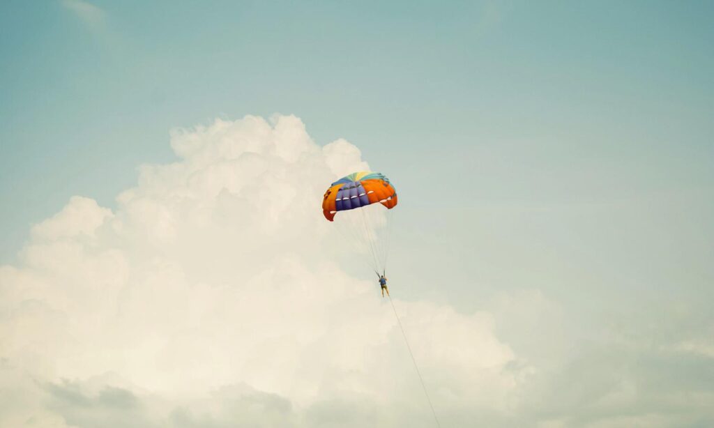 A stylized photo of a parasailor suspended beneath a bright multicolored parachute. He drifts against towering summer clouds, capturing the adventurous coastal recreation and skyborne views that define Florida’s beaches and continue to inspire writers exploring Volusia County’s landscape and creative culture..